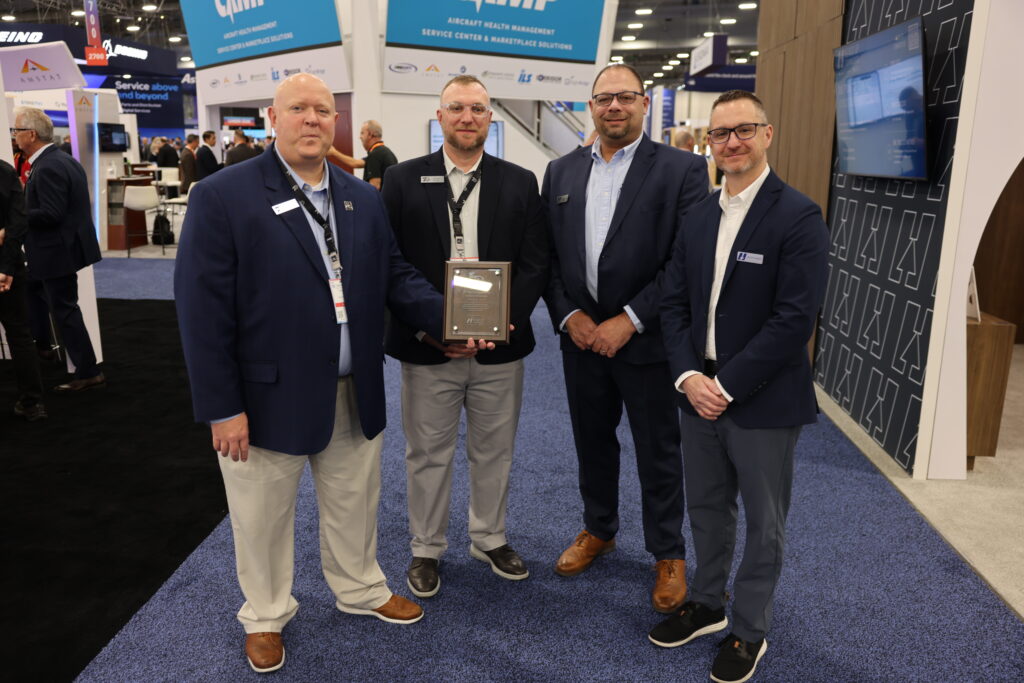 Four men in navy blazers stand together near the Heads Up Technologies booth at an aviation trade show, two of them holding a framed commemorative plaque