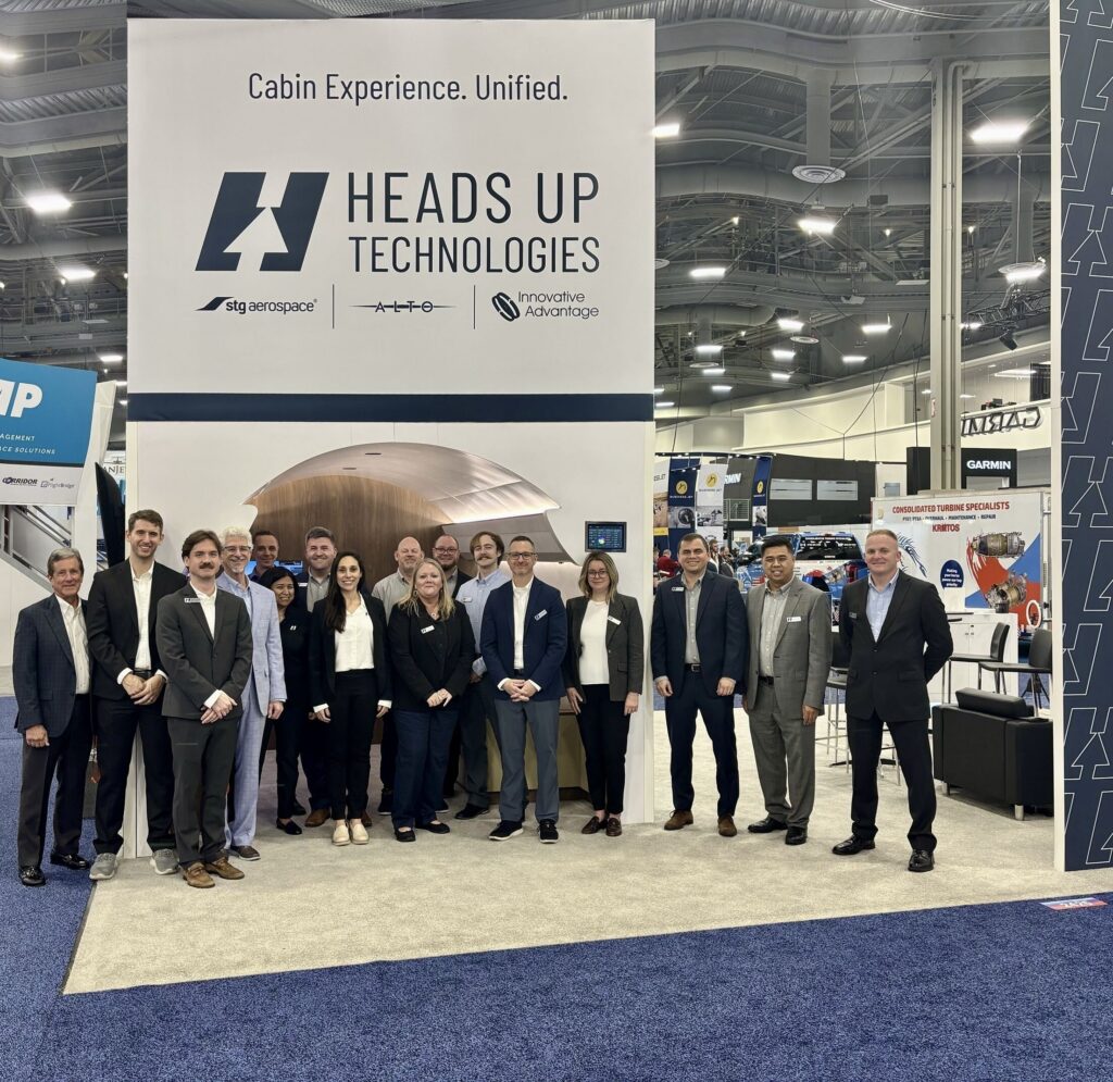 Heads Up Technologies team members in business attire standing in front of their trade show booth, which features a “Cabin Experience. Unified.” banner, STG Aerospace, ALTO, and Innovative Advantage logos, and an aircraft cabin display.